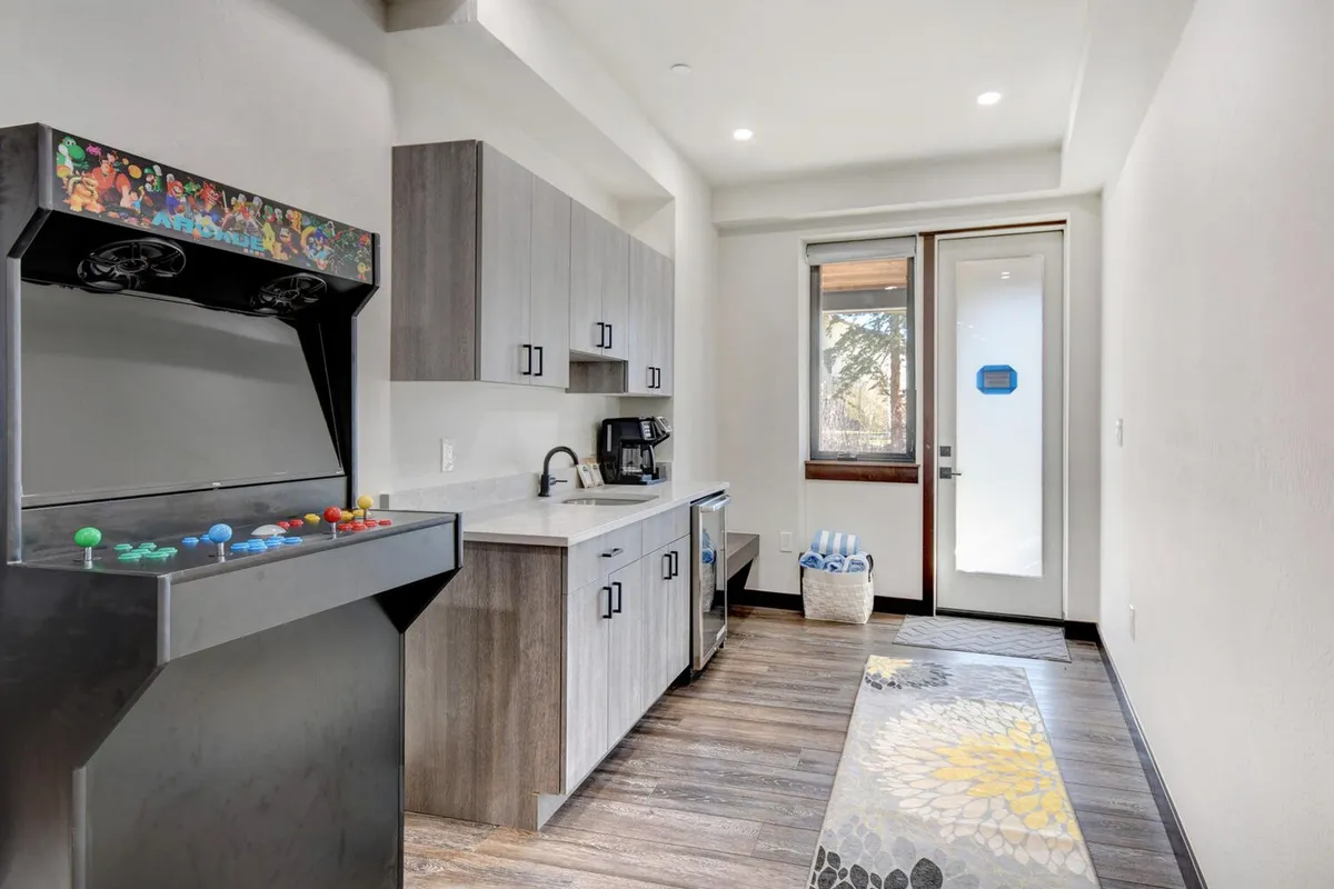 A playful, modern interior showcasing an arcade game positioned near sleek cabinetry and a large window, with a kitchen countertop visible in the foreground. The atmosphere is vibrant and welcoming, perfect for group activities and family fun.