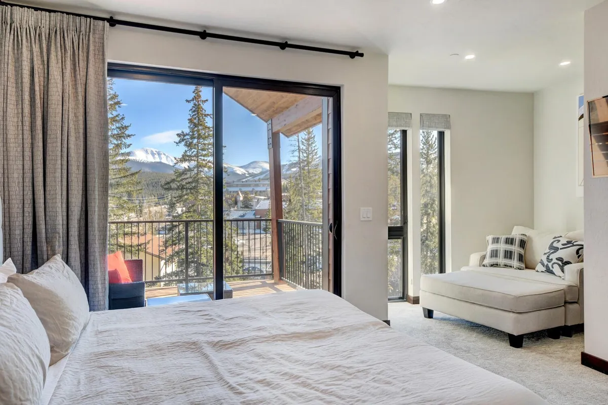 A serene master bedroom with white linens, a large glass door opening to a balcony overlooking mountain views, and a comfortable chair in the corner. The room is flooded with natural light, creating a peaceful retreat.