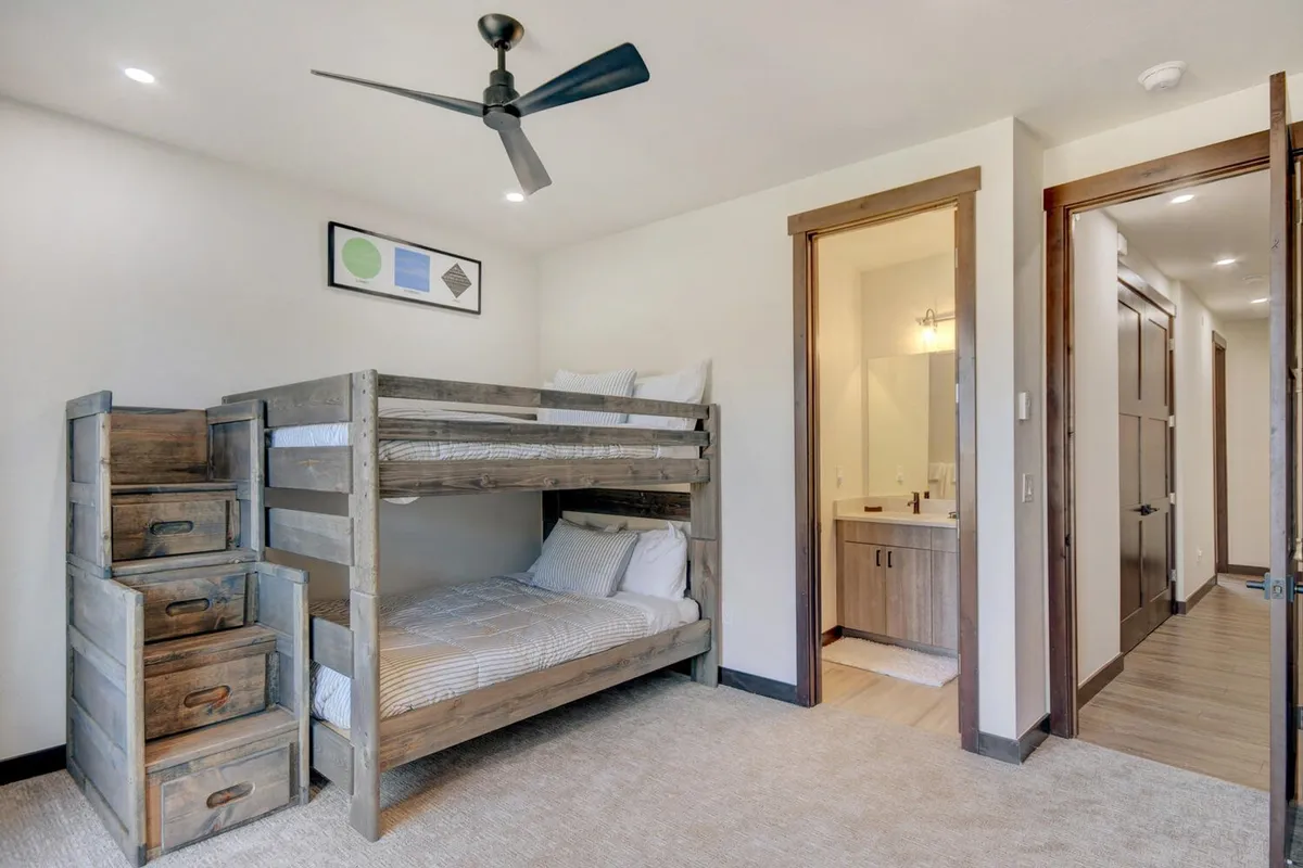 A cozy bunkroom featuring multiple bunk beds, wooden storage drawers, a ceiling fan, and playful framed artwork. The room is designed for comfort and shared experiences, making it perfect for kids or groups.