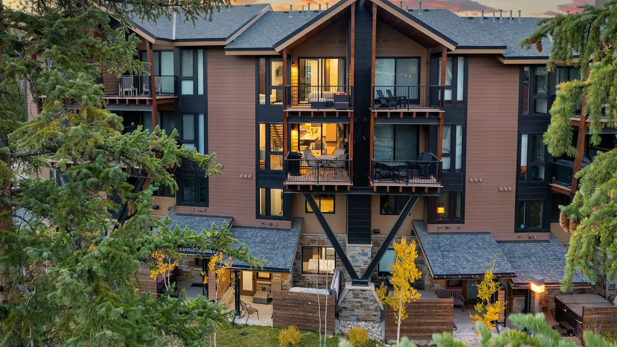 The image showcases the exterior of a luxurious single-family villa set amidst tall trees, with expansive balconies and large windows that capture the surrounding mountain scenery. The scene is warmly lit by decorative outdoor lighting, and a stone pathway invites guests toward the entrance. The tranquil ambiance is accentuated by the promise of sunset views from the balcony and the cozy glow of an outdoor fire pit area, perfect for relaxing or gathering after a day of adventure.