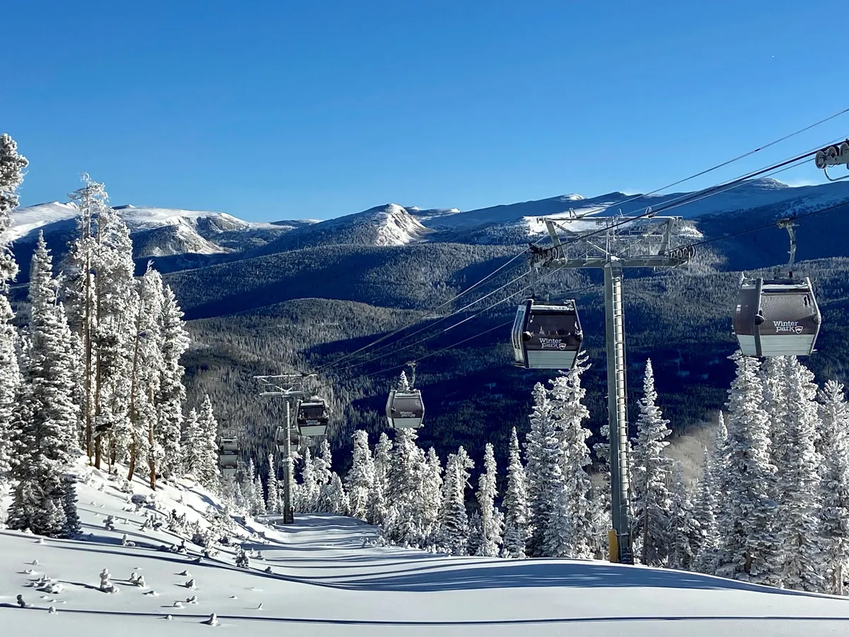 This image captures ski lift gondolas gliding above snow-covered trees with a majestic mountain range in the background. The snow-laden landscape suggests an active winter sports environment, with the lifts providing direct access to the slopes.