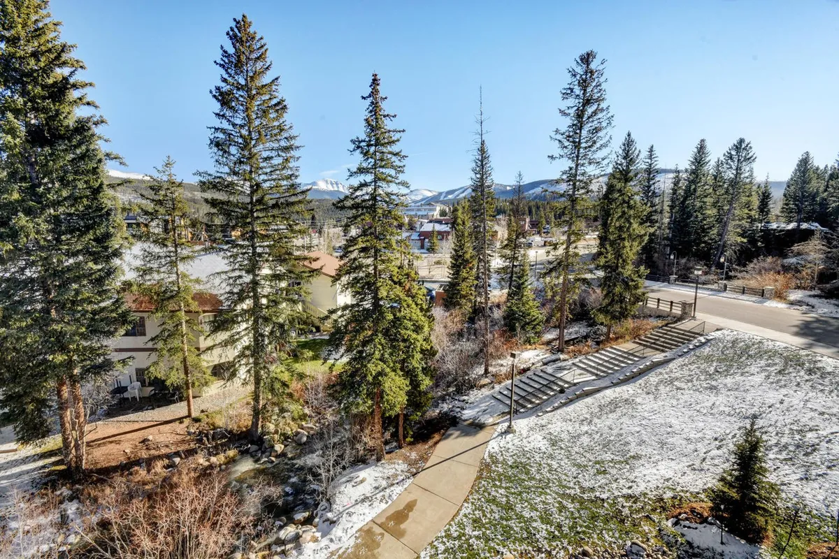 A tranquil pathway meanders through tall pine trees and a snow-covered landscape, with a majestic mountain range in the background. The scene is peaceful, inviting exploration and quiet reflection in nature.