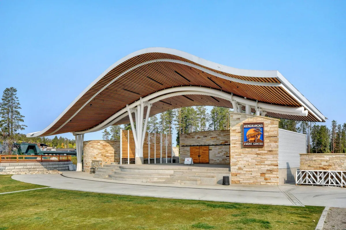 The image features the iconic Hideaway Park Amphitheater with its distinctive curved roof, stone facade, and event signage. The scene evokes a sense of community with hints of outdoor events and concerts, set against a backdrop of mountain town architecture.