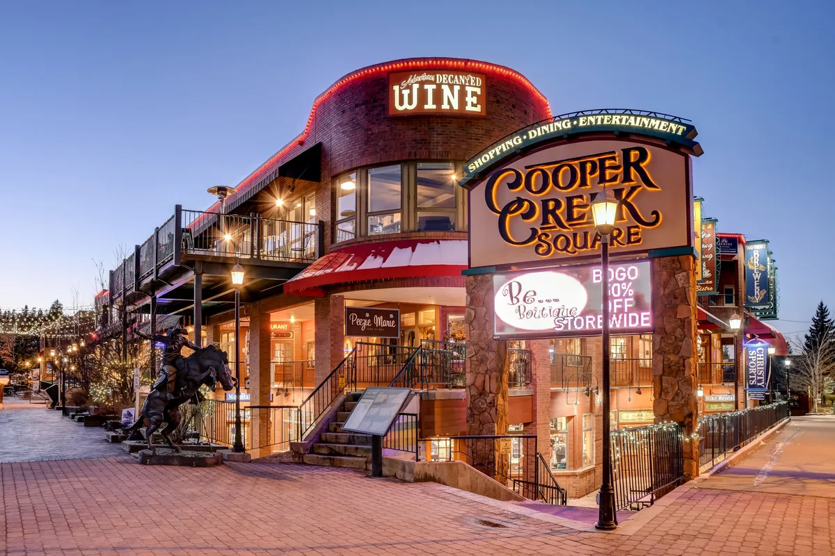 This image shows the lively Cooper Creek Square in downtown Winter Park, featuring decorative lighting, outdoor seating, signage for shops and a wine store, and an artistic sculpture. The street-level view captures a bustling hub for shopping, dining, and entertainment in the town's core.