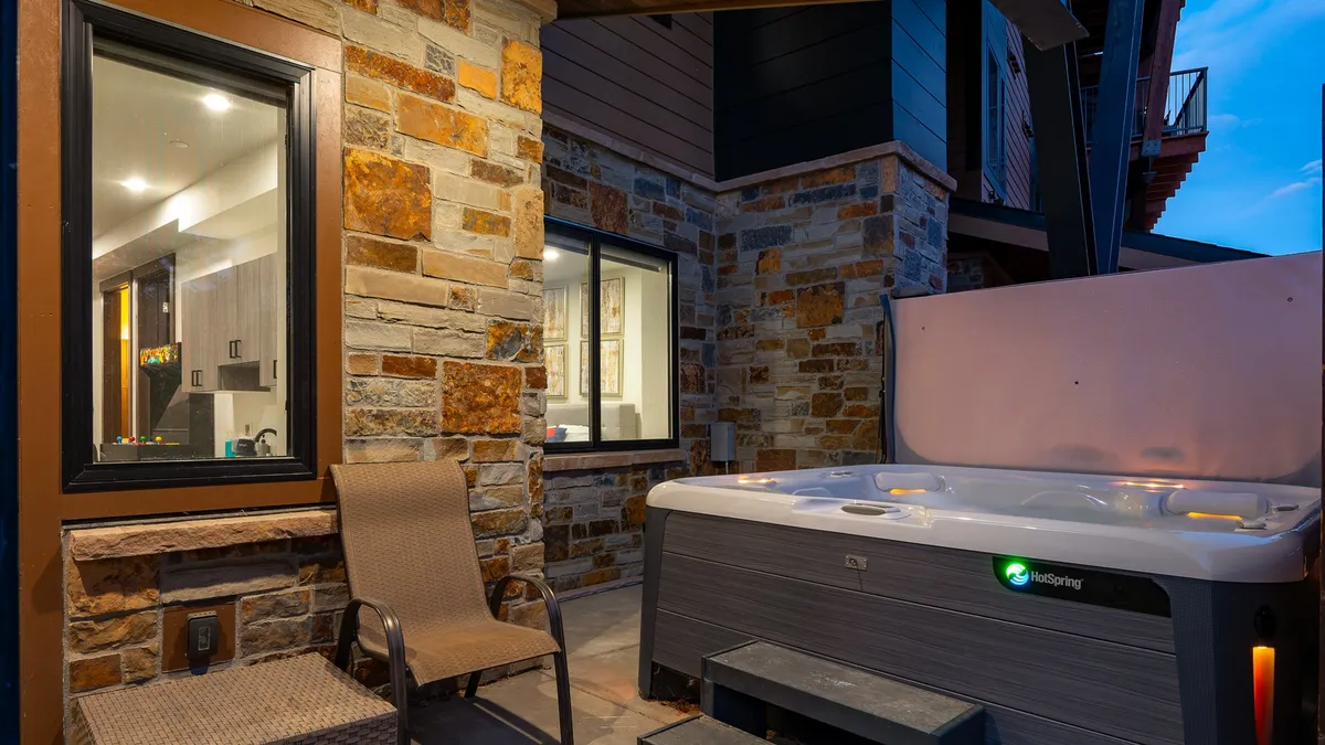 The image shows a tranquil outdoor area featuring a private hot tub nestled against a stone wall, complemented by comfortable outdoor seating. The ambiance is cozy, with the hot tub inviting guests to soak as evening sets in, suggesting opportunities for relaxation and stargazing.