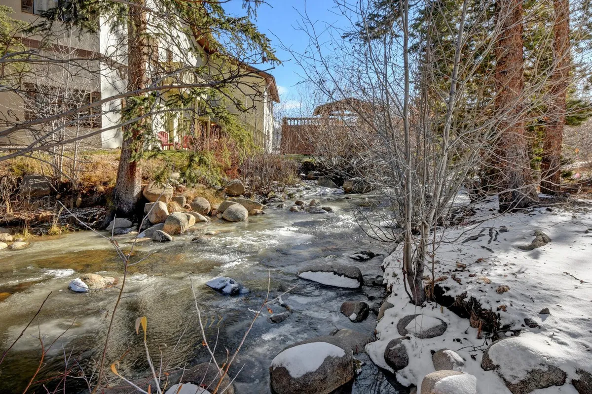 A peaceful scene featuring a flowing creek bordered by rocks and trees with the property in the background. The stream is the clear focal point, with an atmosphere of quiet connection to nature and the sounds of running water.