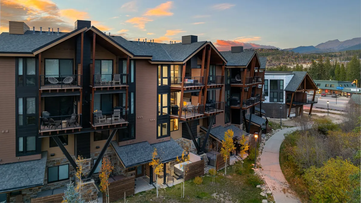 A welcoming exterior shot of a modern mountain property with balconies, surrounding trees, and a pathway leading toward the town. The image gives a sense of easy access to both natural beauty and urban amenities, with the architecture blending into the lively, walkable environment.