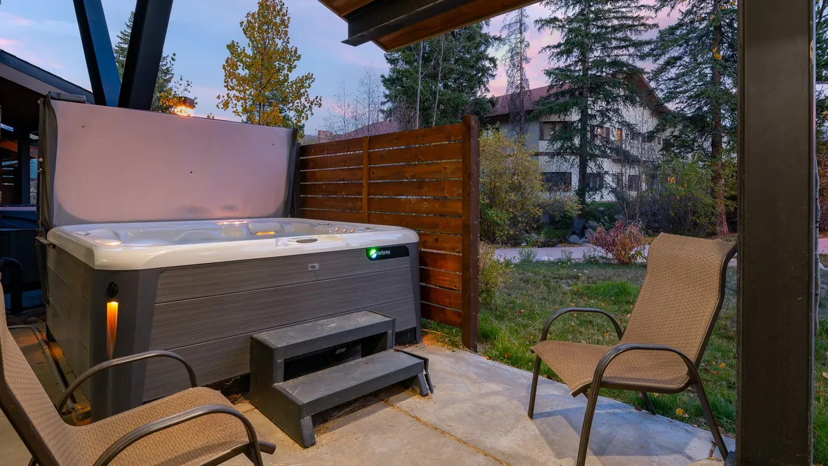 This image captures a tranquil outdoor hot tub area surrounded by a wooden fence and outdoor seating, set against the backdrop of treetops as the sun sets. The setting is inviting, with the hot tub as the central feature, suggesting warmth and relaxation amid the natural landscape.
