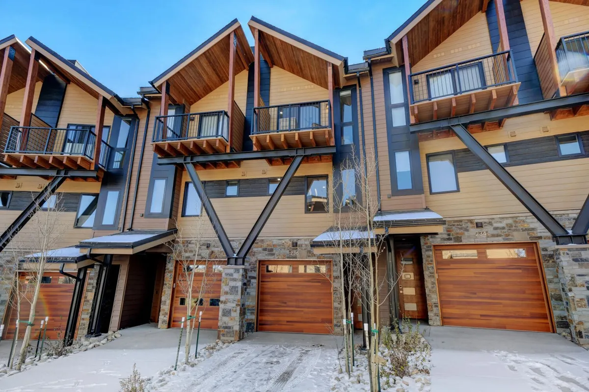 A welcoming exterior view of the villa, showcasing its wooden balconies, stone façade, large windows, and garage doors. The scene hints at both wintertime and summer activities, with inviting outdoor spaces and convenient access.