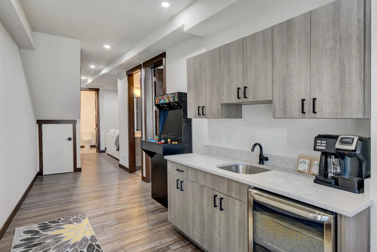 A stylish open-plan room with crisp modern cabinetry, kitchen appliances, an arcade game, and a decorative rug. This multi-functional space is set up for both dining/entertaining and energetic game nights, blending relaxation and excitement.